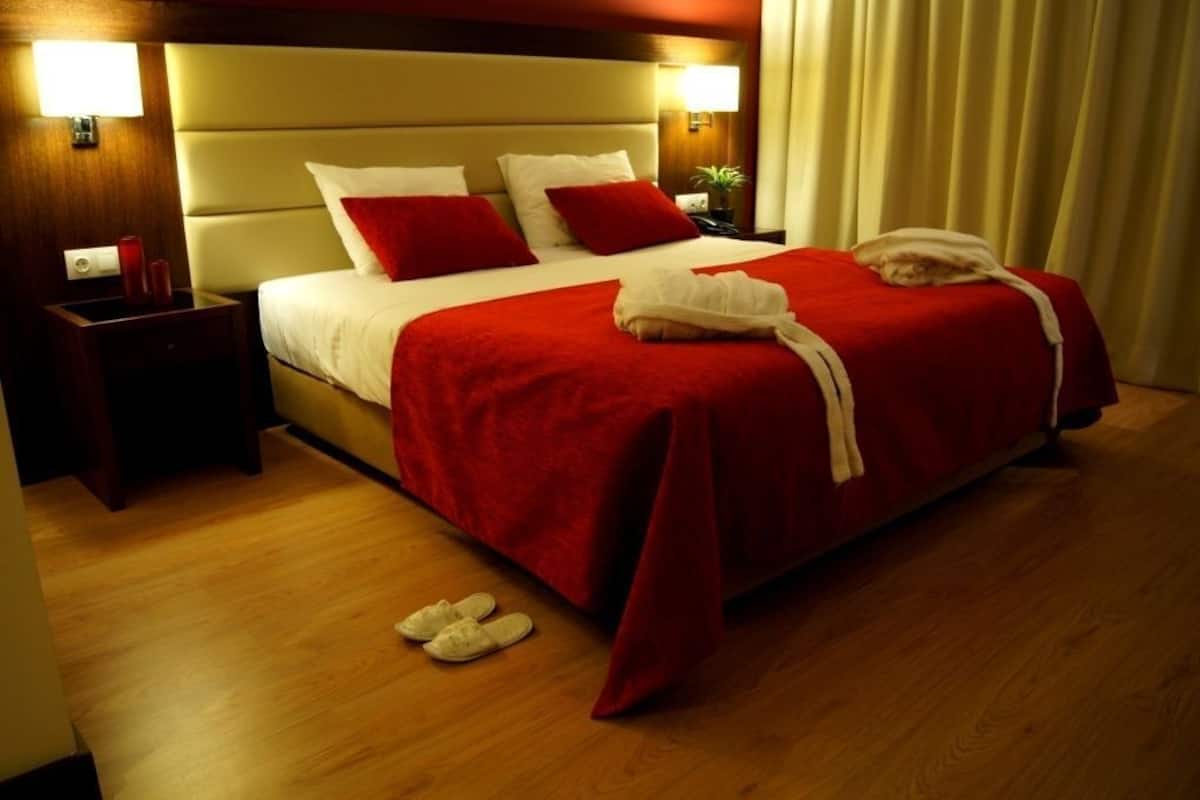 Une chambre d’hôtel avec un grand lit, deux tables de chevet, une petite table, une couverture rouge et une serviette blanche sur le lit.