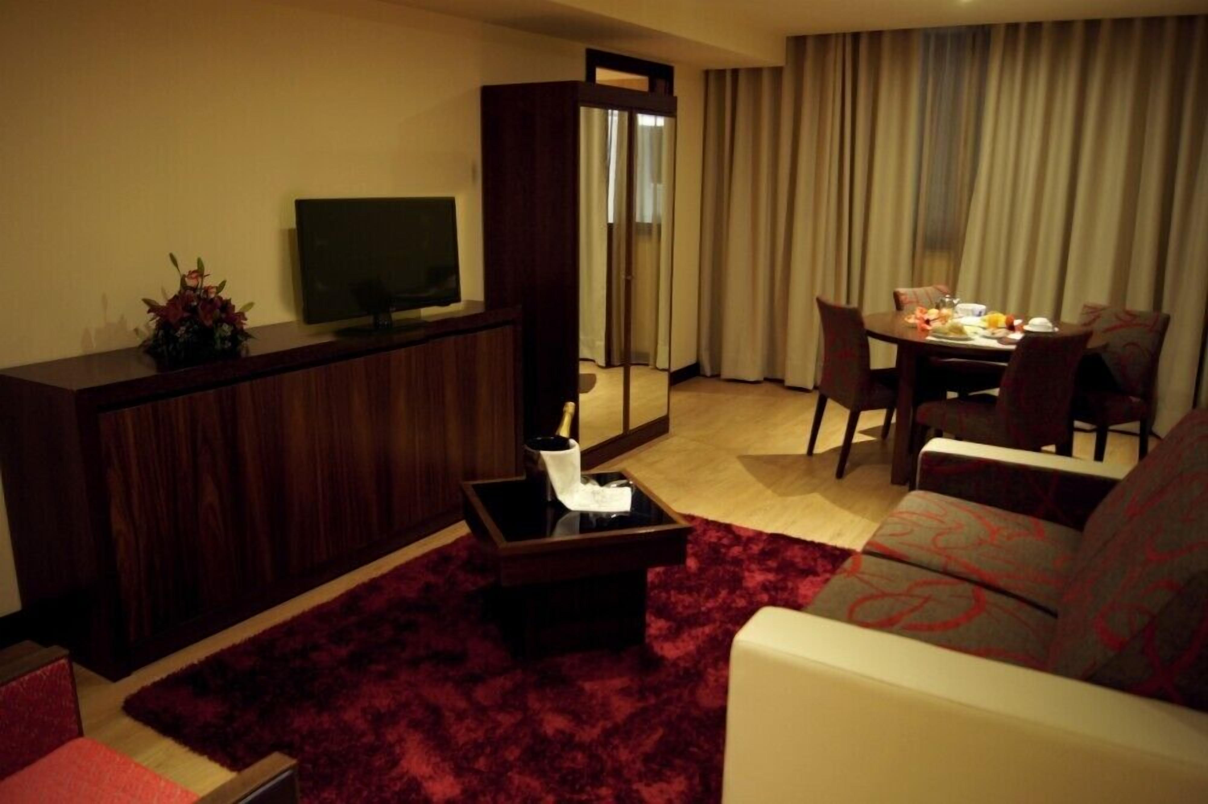 A modern living room with a dark wood cabinet, a flat-screen TV, a red carpet, a sofa, and a dining area with a table set for two.