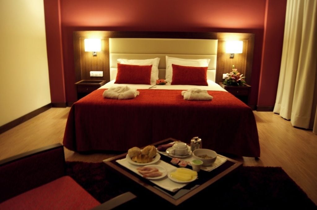 Habitación de hotel con una cama grande, dos lámparas de noche, una bandeja con alimentos para el desayuno y una mesita de noche con lámpara y flores.