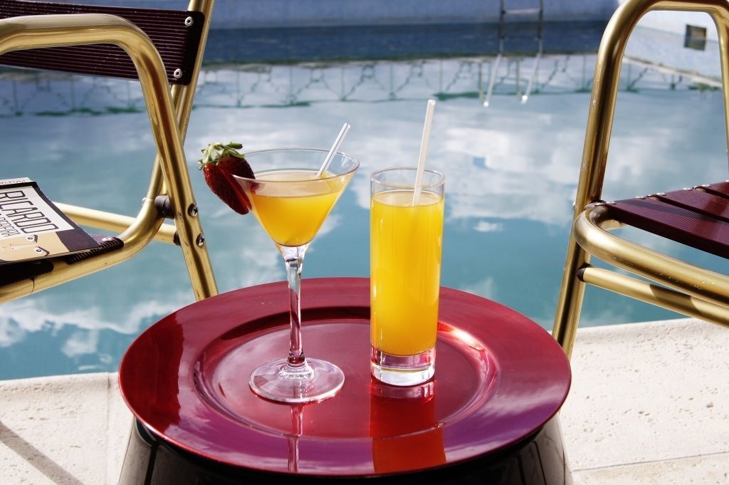 Une table sur laquelle se trouve un plateau rouge, avec un verre à martini et un grand verre de jus d’orange. On aperçoit une piscine et le ciel en arrière-plan.