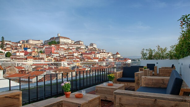 A rooftop terrace with wooden furniture, potted plants, and a view of a cityscape with red-roofed buildings. - Hotel Oslo (Coimbra)