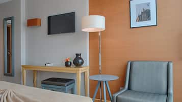 Habitación de hotel con cama, escritorio con silla, lámpara de pie, televisión y un cuadro en la pared.