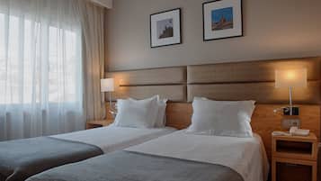Et hotelværelse med to senge, et stort vindue med lette gardiner, to billeder hængende på væggen og sengeborde med lamper.