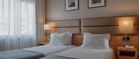 A hotel room with two beds, a large window with sheer curtains, two framed pictures on the wall, and bedside lamps.