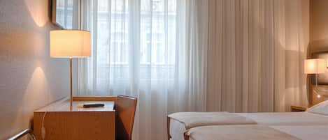 A hotel room with a desk, chair, two beds, a lamp, and a large window with sheer curtains.