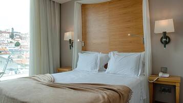 Quarto de hotel com uma cama grande, duas luminárias de cabeceira, uma cabeceira de madeira, uma mesa de cabeceira com telefone e vista para a cidade.
