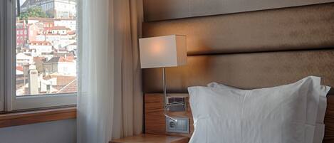 A hotel room with a bed, a nightstand, a lamp, and a view of a cityscape through a window.