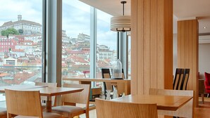 A modern dining area with large windows offering a city view, wooden furniture, and a pendant light. - Hotel Oslo (Coimbra)