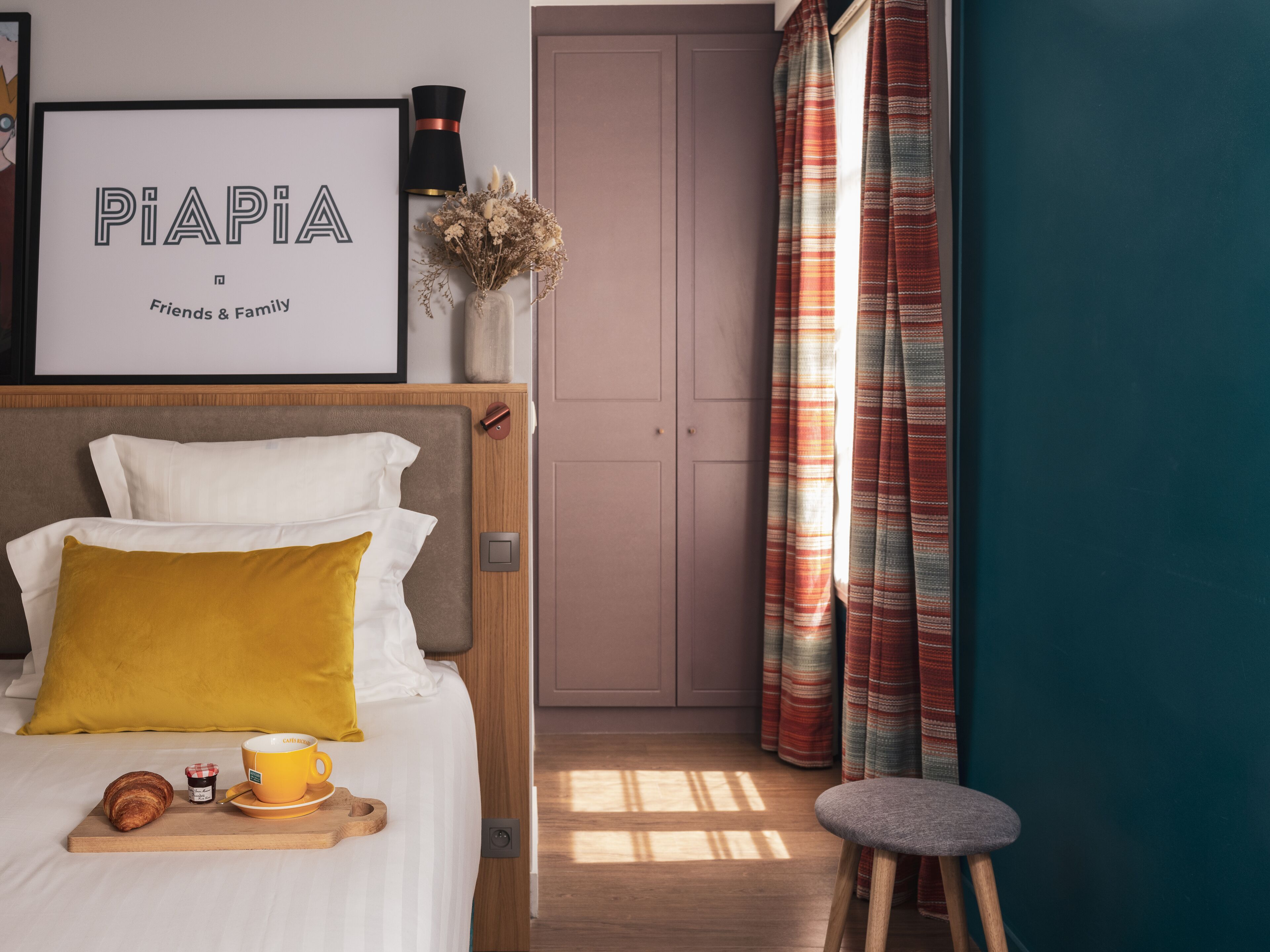 Una cama bien hecha con un almohadón amarillo, un cartel enmarcado encima, una mesita con una taza y un croissant, y un taburete.