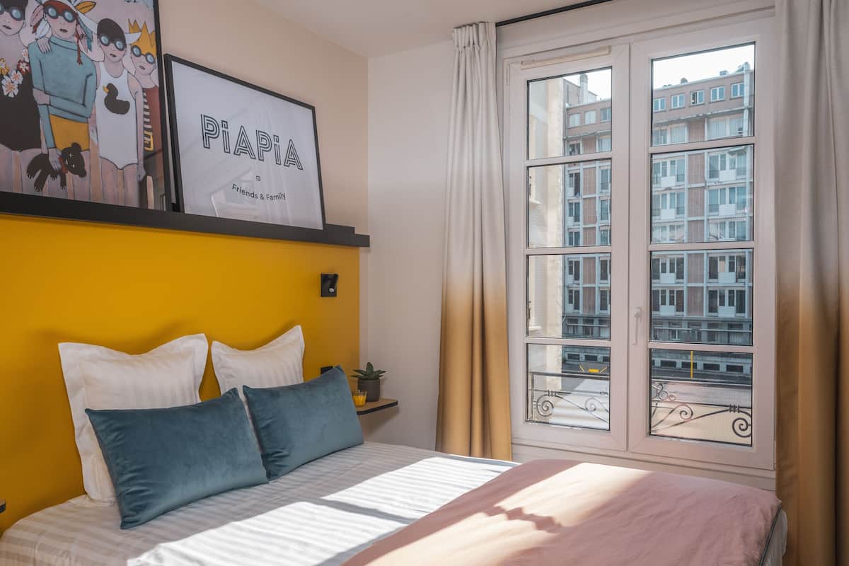 Une chambre à coucher avec un lit, des oreillers, une table de chevet, un tableau encadré et une fenêtre donnant sur des immeubles.