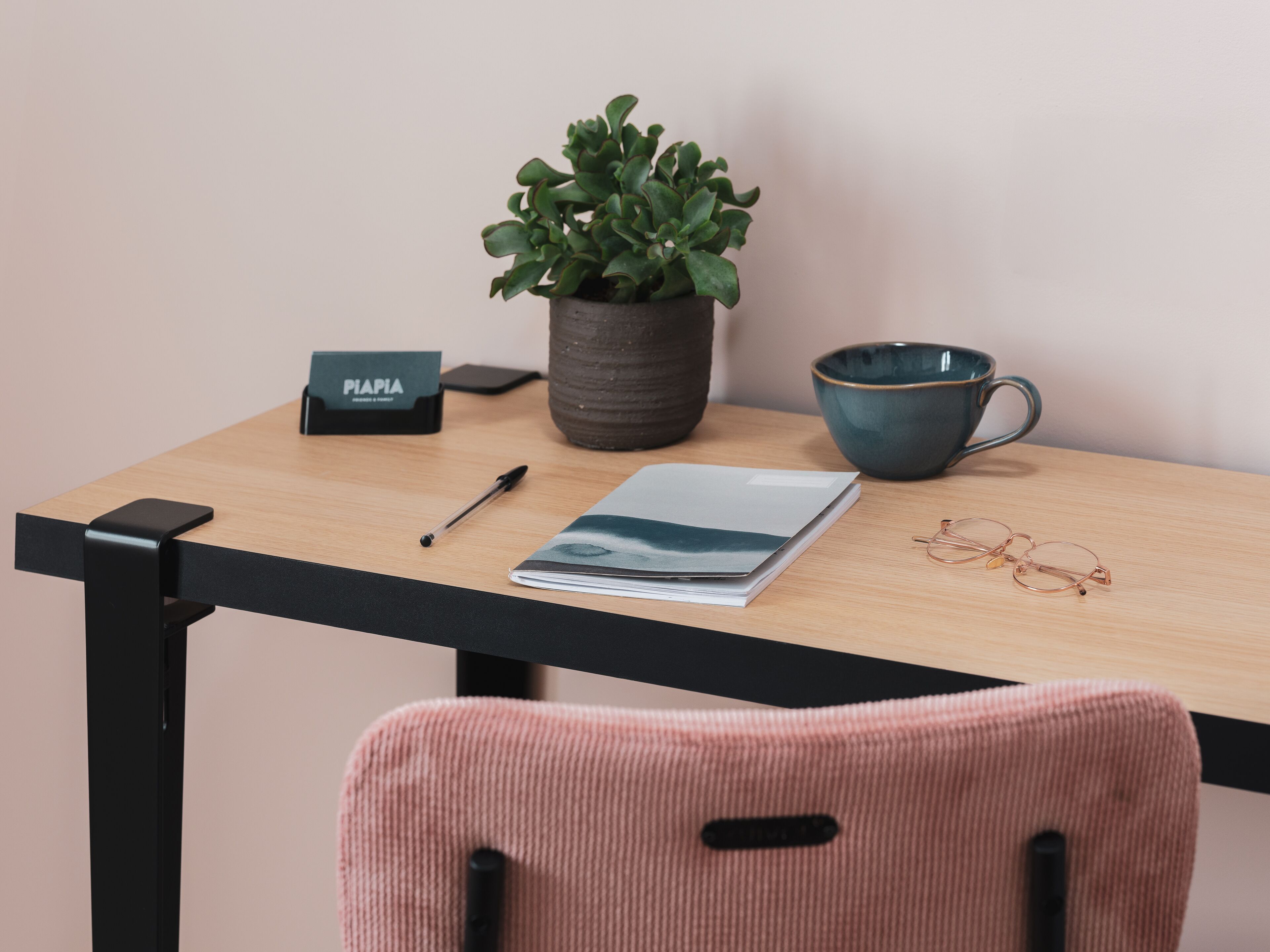 A wooden desk with a potted plant, a notebook, a pen, a pair of glasses, a ceramic mug, and a small sign.