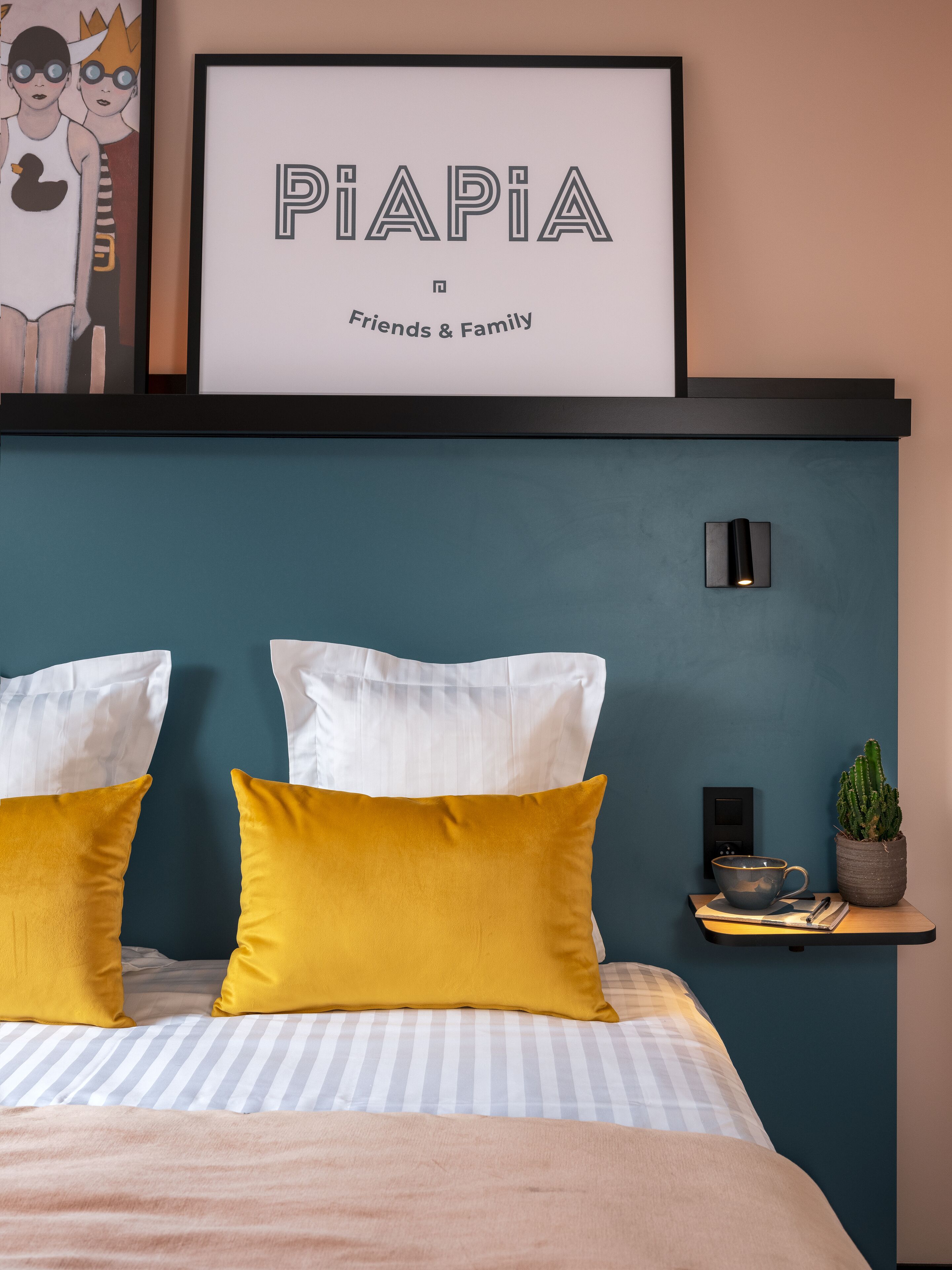 Un dormitorio con cama, almohadas, una mesita de noche, una obra de arte enmarcada y una lámpara de pared.