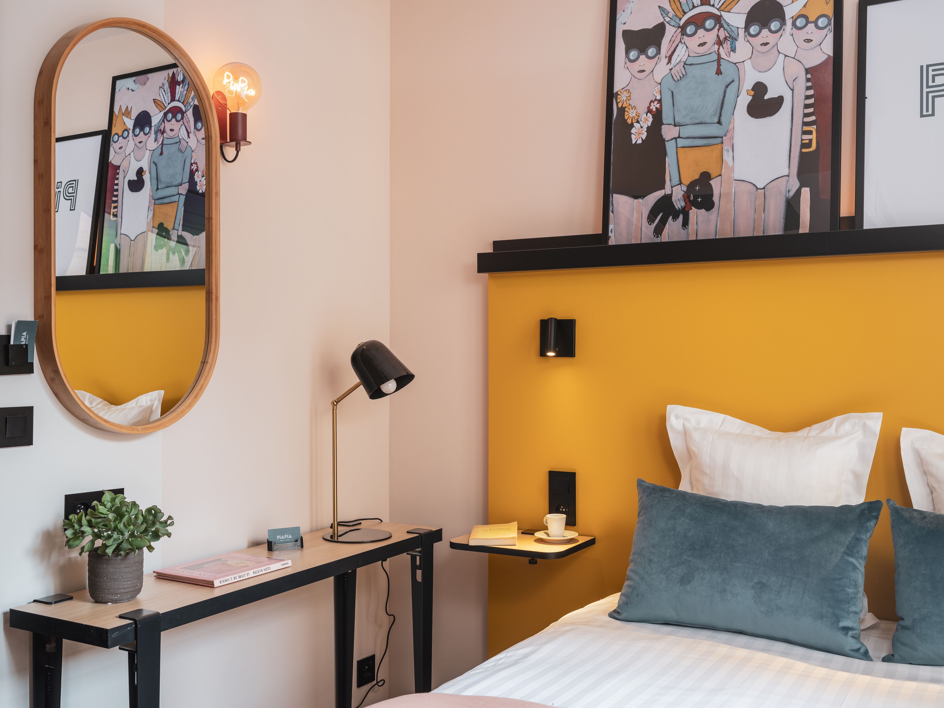 A hotel room with a bed, a bedside table with a lamp, a mirror, and artwork on the wall.