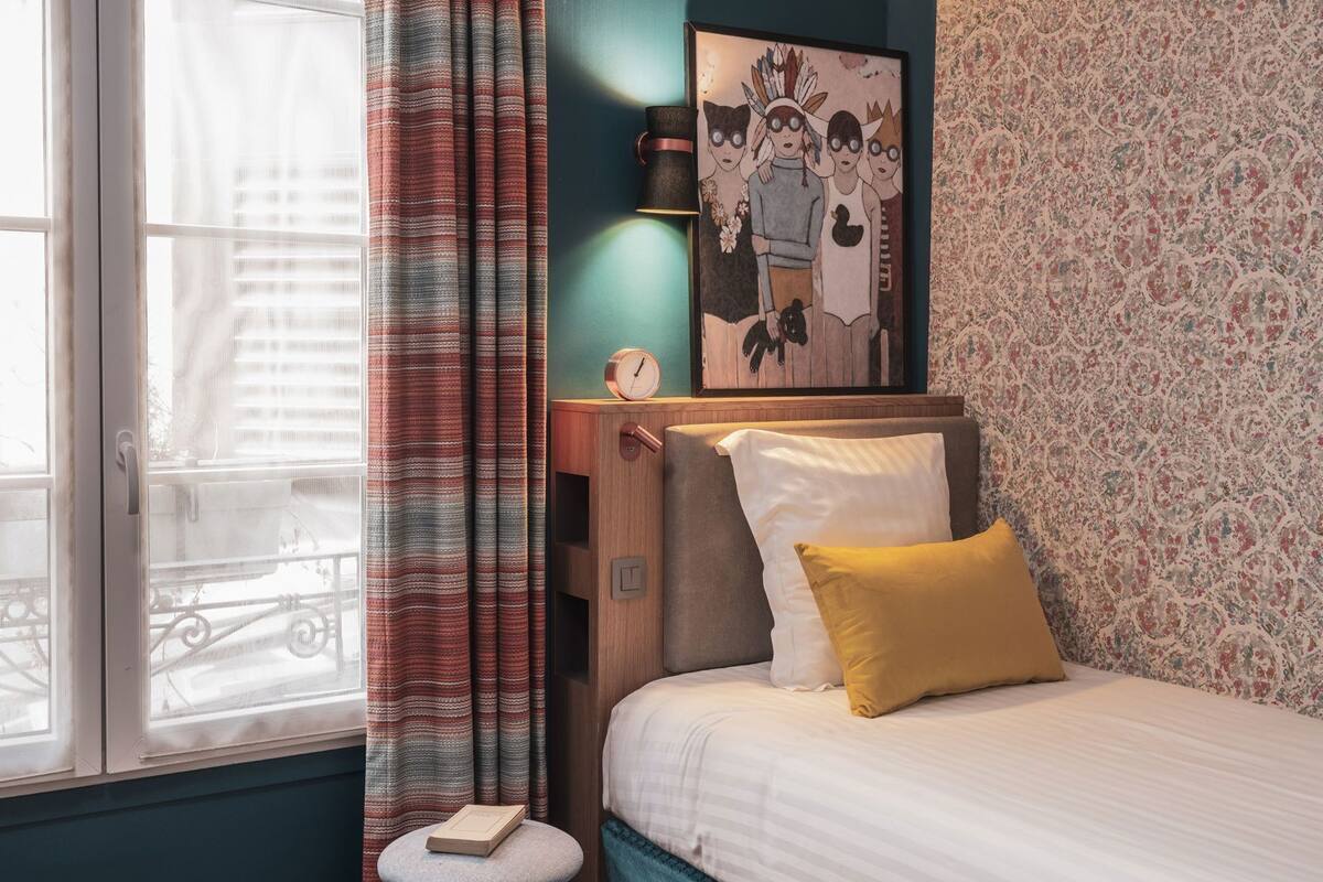 Une chambre d’hôtel avec un lit, un petit tabouret, une horloge et une fenêtre avec des rideaux.