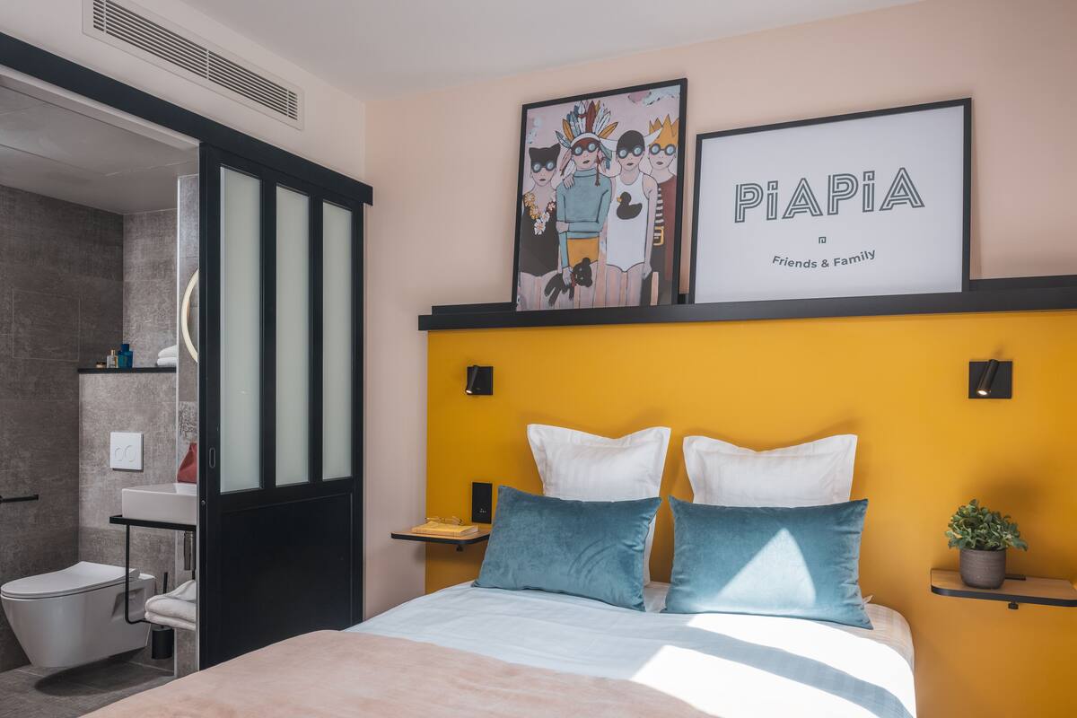 Une chambre à coucher moderne, avec un lit, une table de chevet, une étagère fixée au mur et une salle de bain visible à travers une porte ouverte.