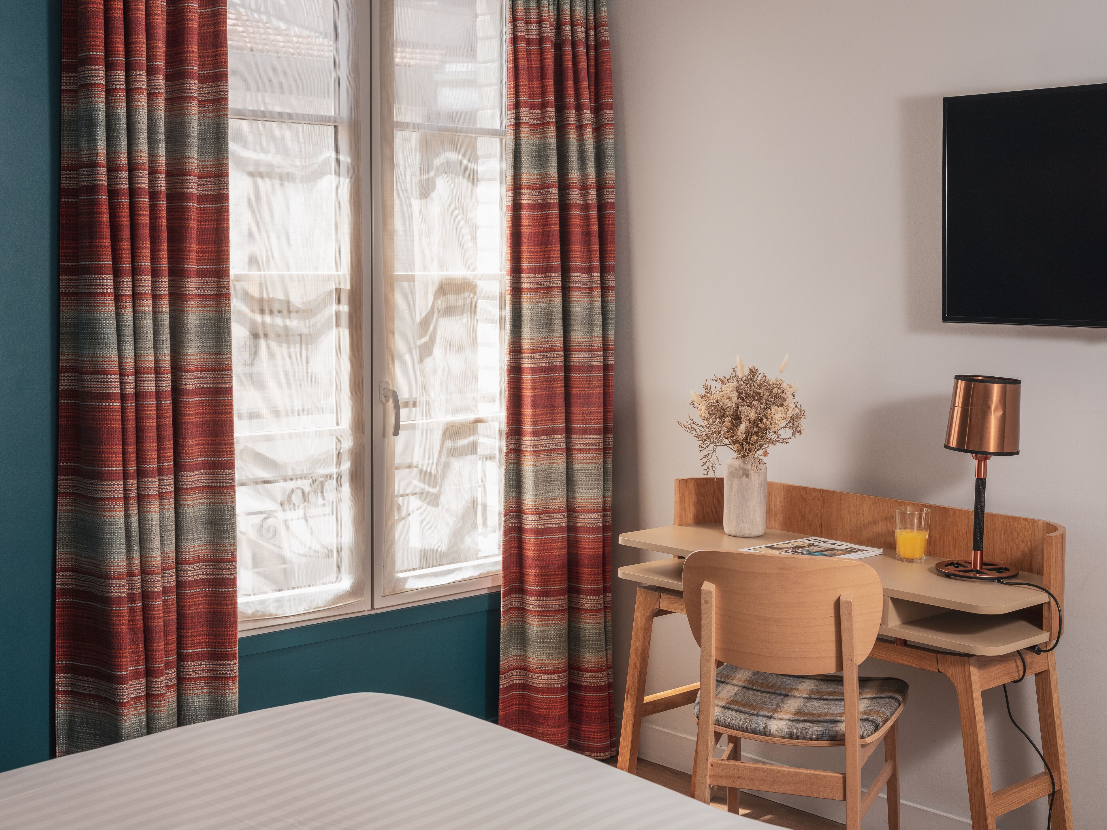 Una habitación de hotel con una cama, un escritorio con una silla, una lámpara y un florero con flores secas.