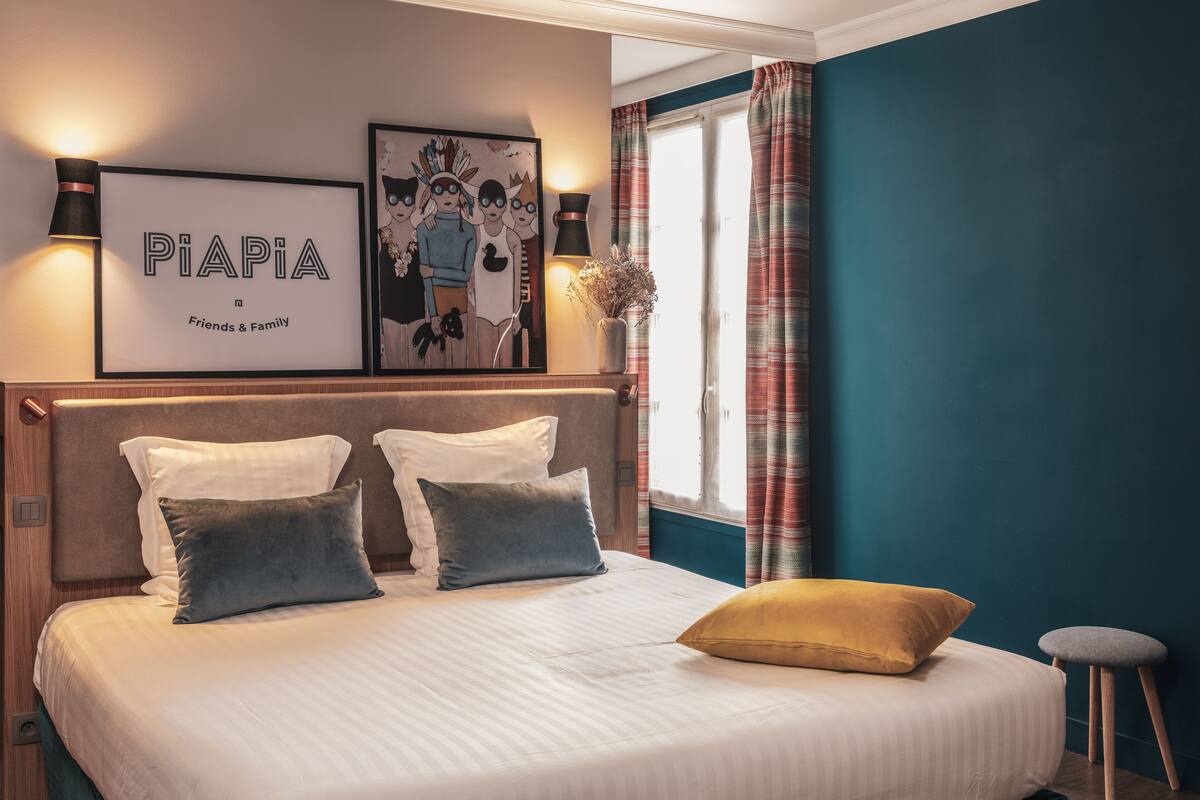Une chambre à coucher comprenant un lit, des oreillers, une table de chevet, une lampe, une fenêtre avec des rideaux et des œuvres d’art mural.