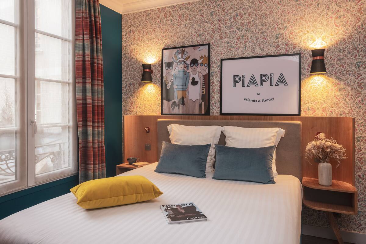 Un lit bien fait, avec un oreiller jaune et un magazine, un mur orné d’un motif floral, et deux tableaux encadrés portant l’inscription « PiAPIA Friends & Family ».