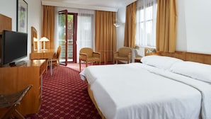 Una habitación de hotel con una cama, un escritorio con televisor, dos sillas, una mesita y una ventana con cortinas.