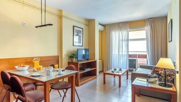 Una habitación de hotel moderna con comedor, zona de estar con sofá y televisión, y ventana con cortinas.