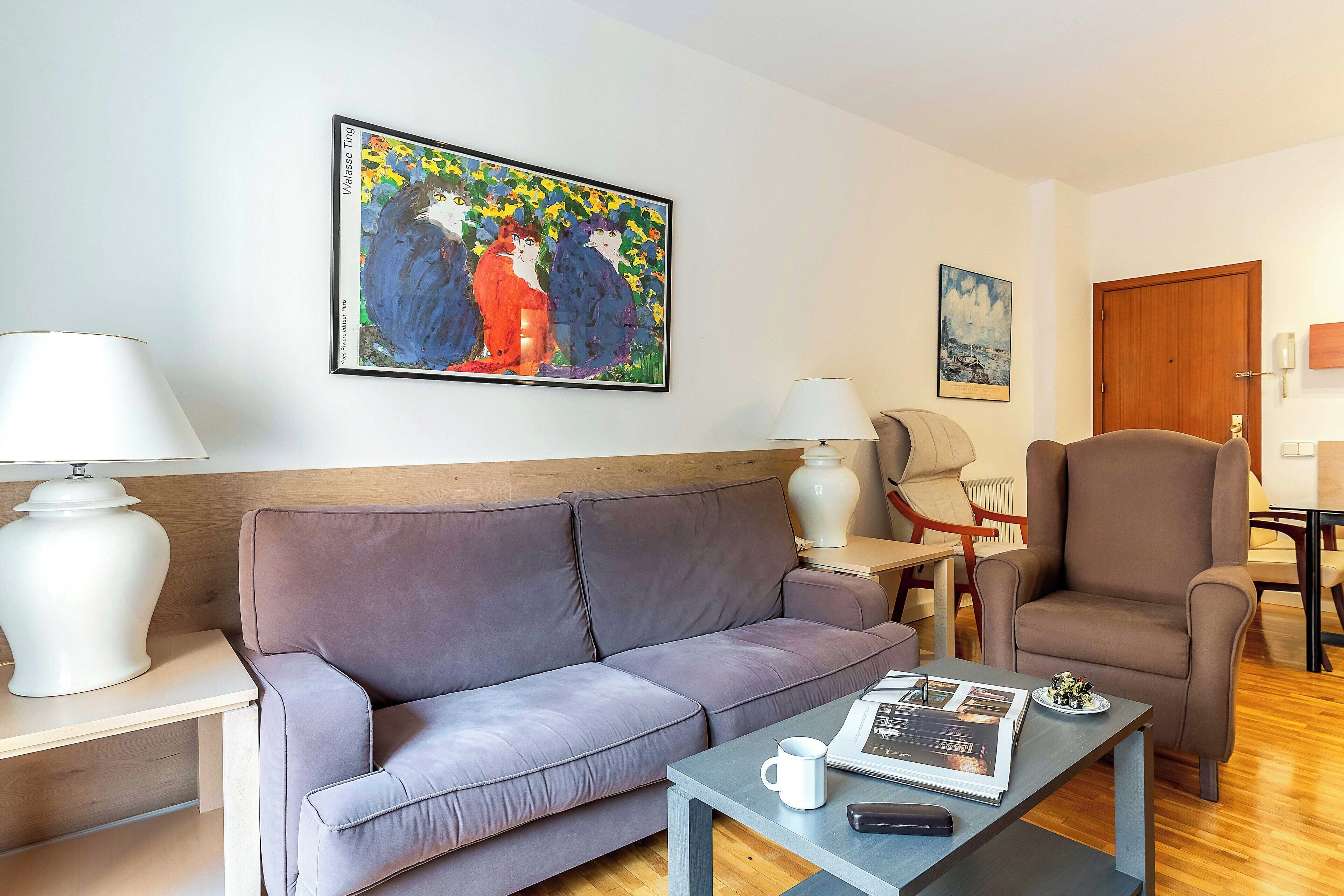 A modern living room with a grey sofa, a coffee table with a remote control, a white lamp, and a painting on the wall.