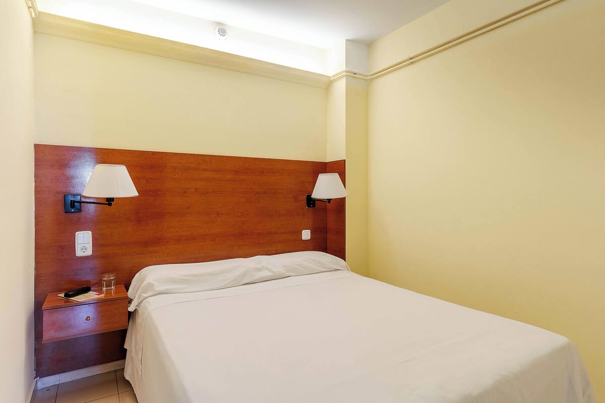 A hotel room with a wooden headboard, two bedside lamps, a bedside table with a glass of water, and a neatly made bed.