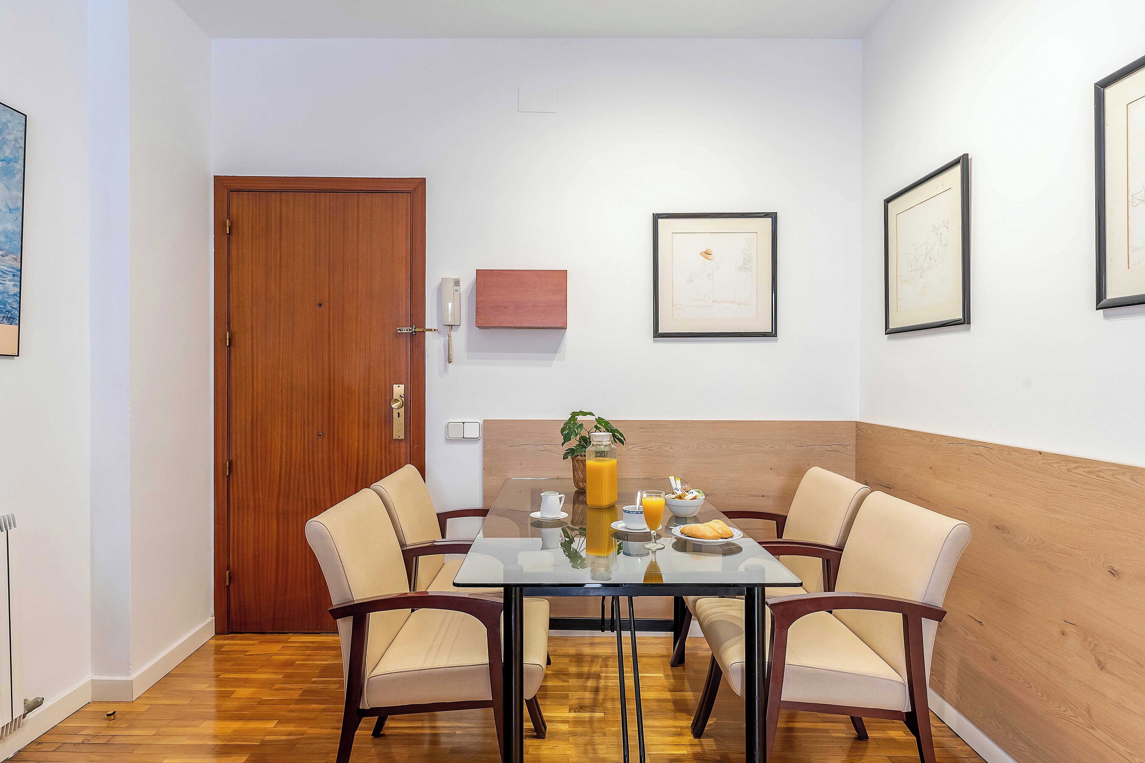 A dining area with a glass table, four chairs, a wooden door, and framed artwork on the walls.