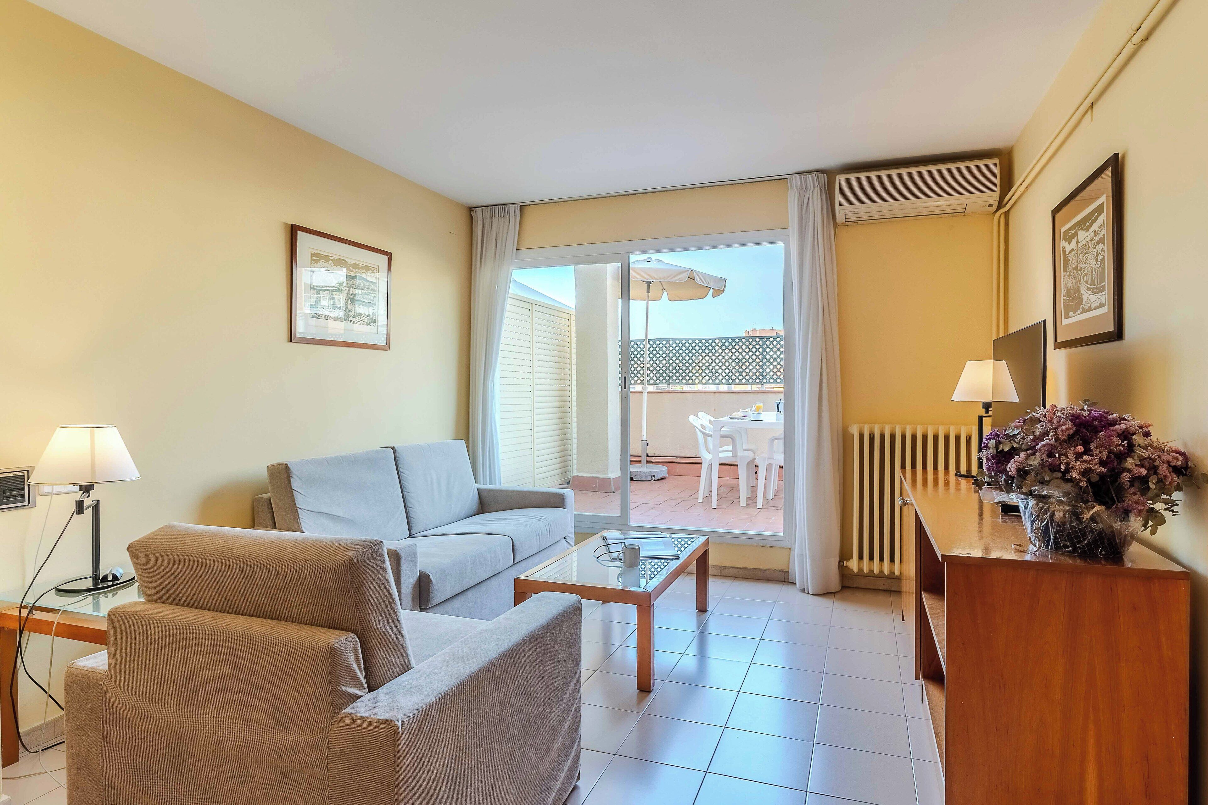 Habitación de hotel con sofá, una mesita de centro, escritorio con lámpara y vista al patio exterior.