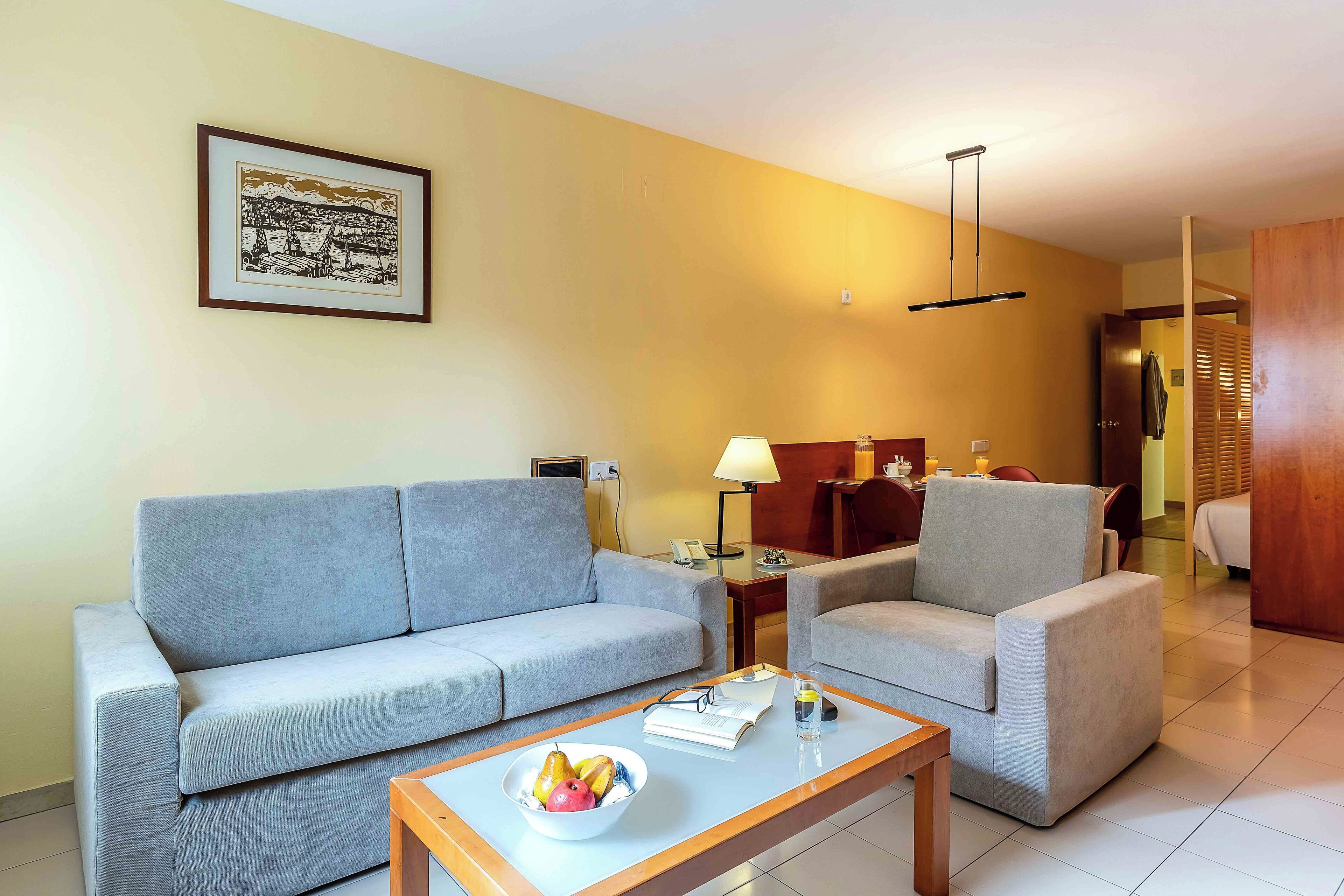 A living room with a sofa, armchair, coffee table with fruit, and a framed picture on the wall.