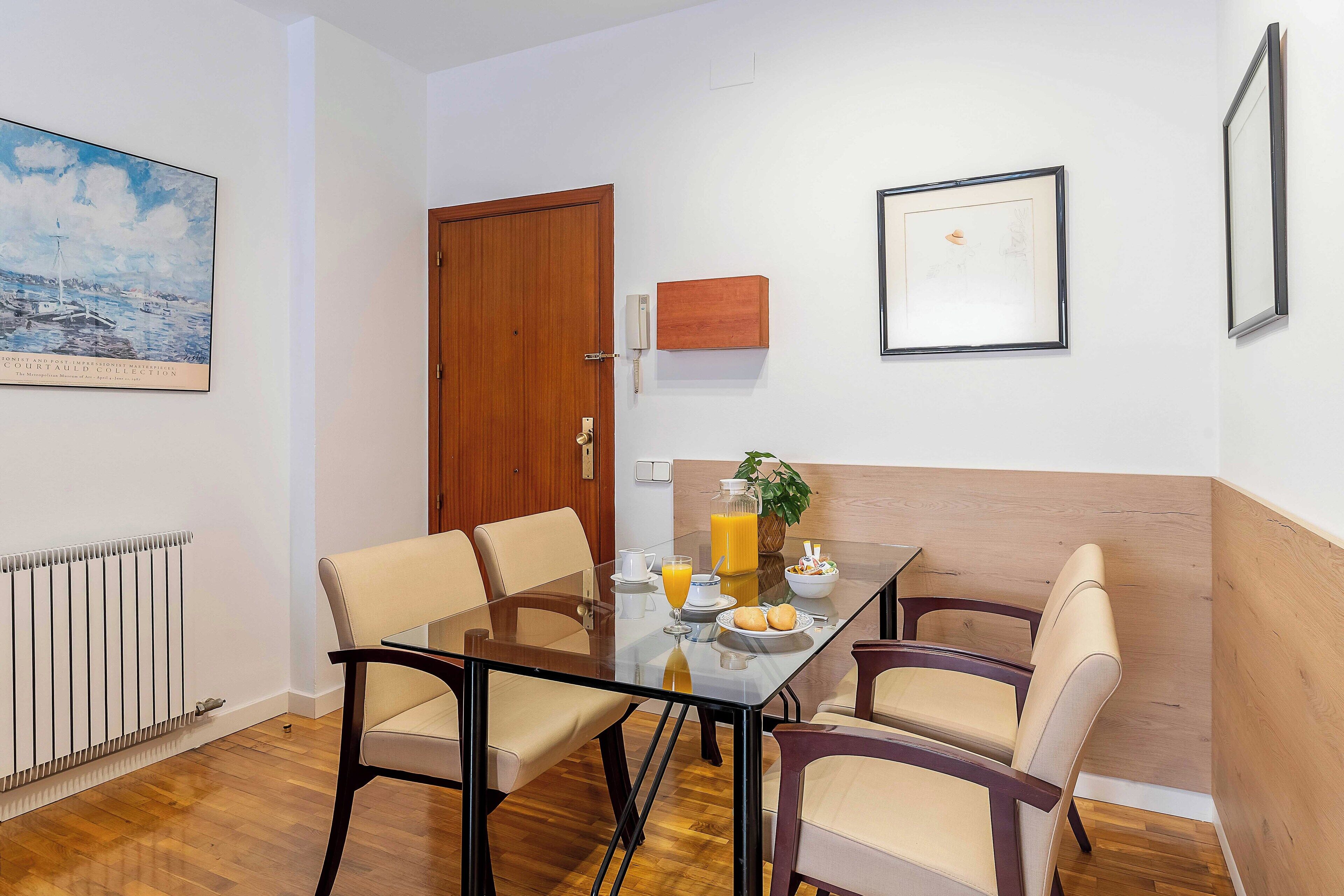 A dining area with a glass table, four chairs, a bowl of fruit, and a bottle of orange juice.