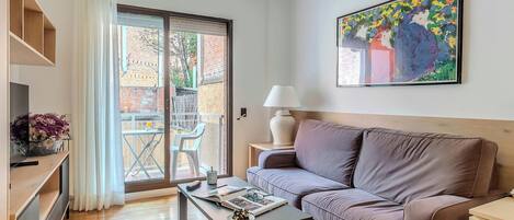 Un salon moderne avec un canapé, une table basse et un téléviseur posé sur un meuble. Une porte-fenêtre coulissante donne accès à un espace extérieur doté d’un balcon.