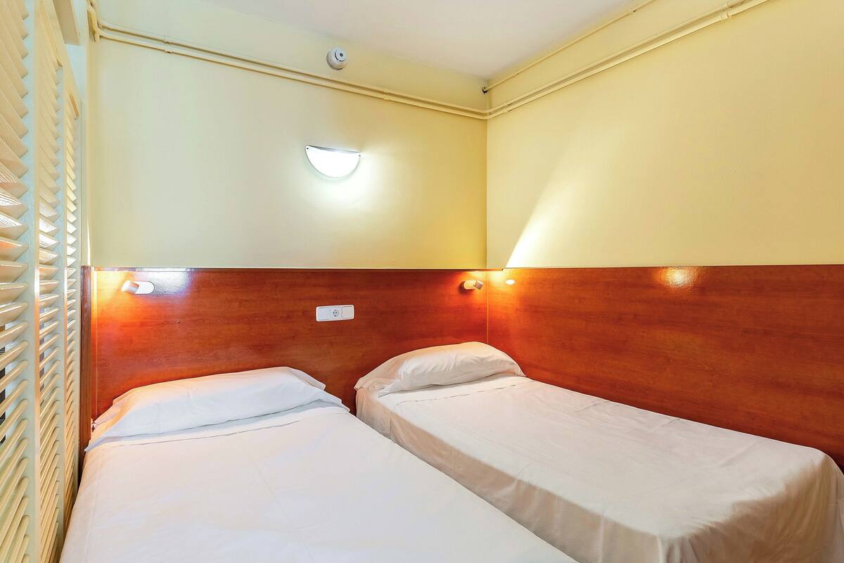 A room with two single beds, a wooden headboard, a wall-mounted light, and a thermostat.