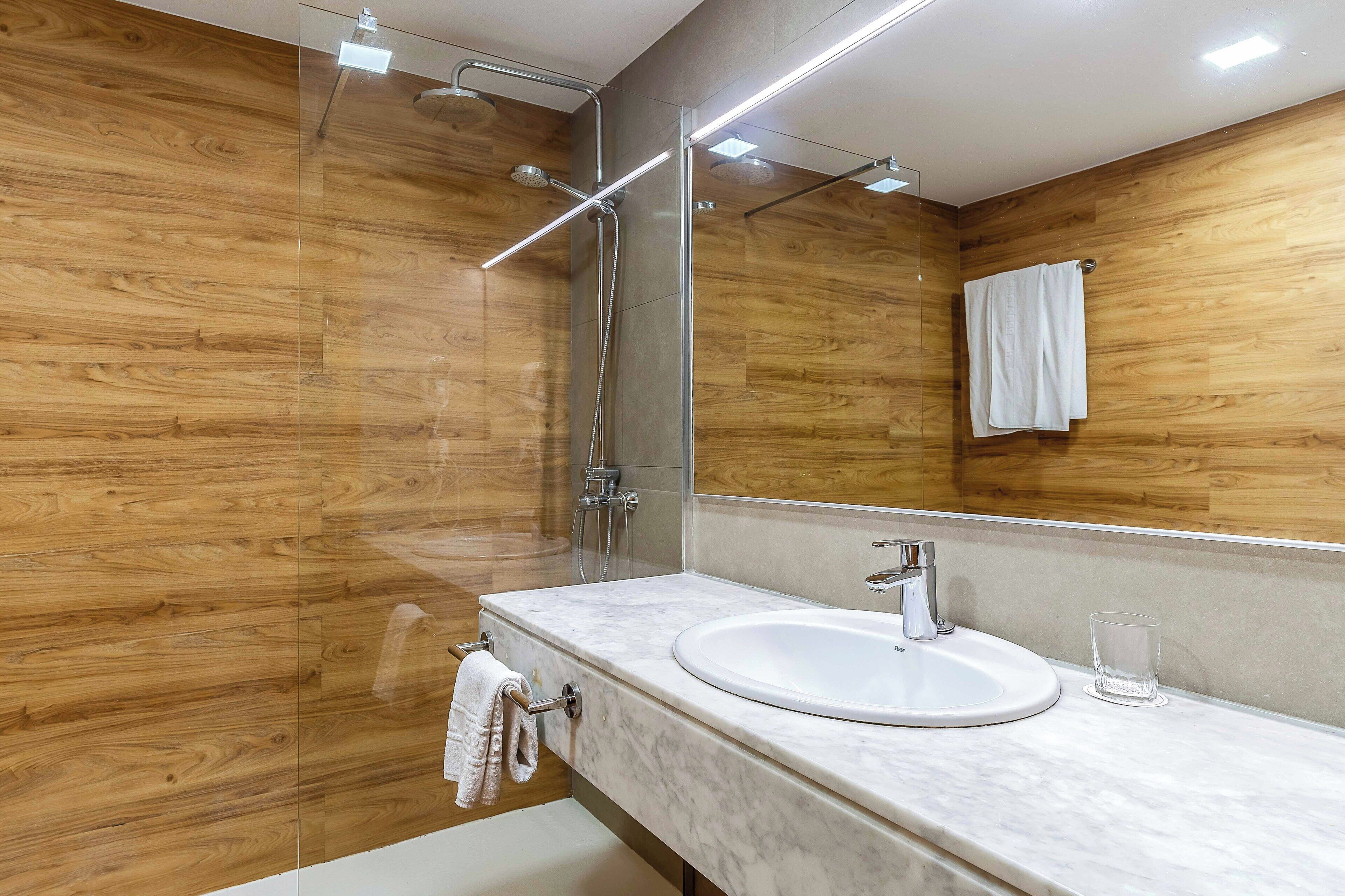 Een moderne badkamer met een houten wand, een glazen douchecabine, een witte wastafel en een glas water op het aanrecht.