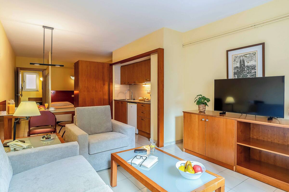 A modern hotel room with a living area, kitchenette, and a TV.