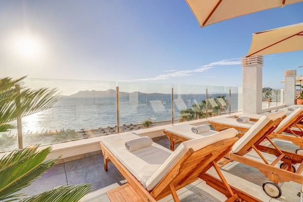Un espace de détente extérieur baigné de soleil, avec des chaises longues en bois, une balustrade en verre et une vue sur la mer.