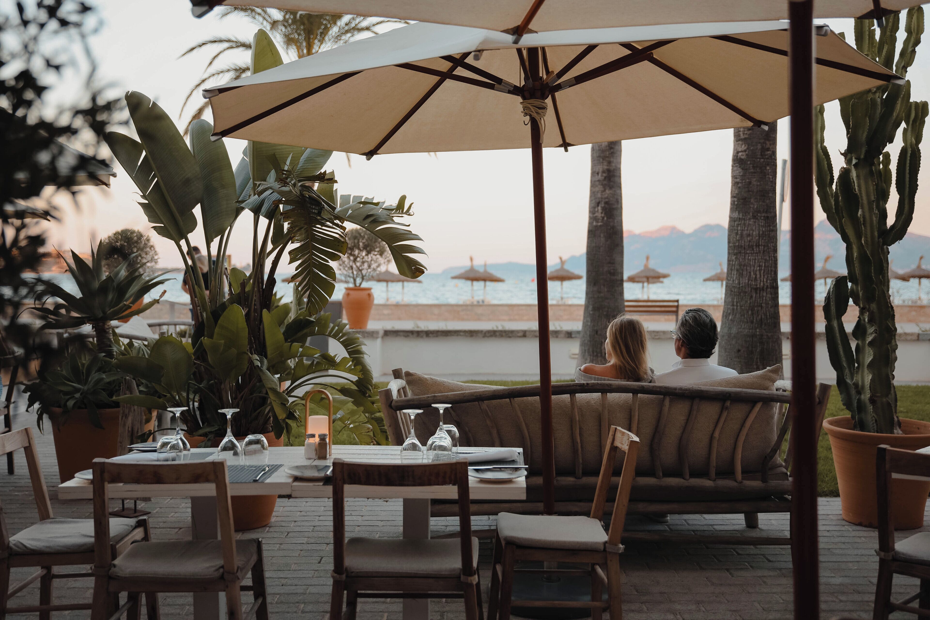 Um casal sentado à mesa com vista para o mar, rodeado por vasos de plantas e guarda-sóis.