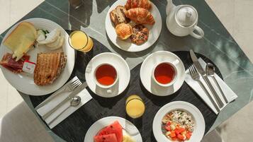 Desayuno con cruasanes, tostadas, fruta y bebidas sobre una mesa.