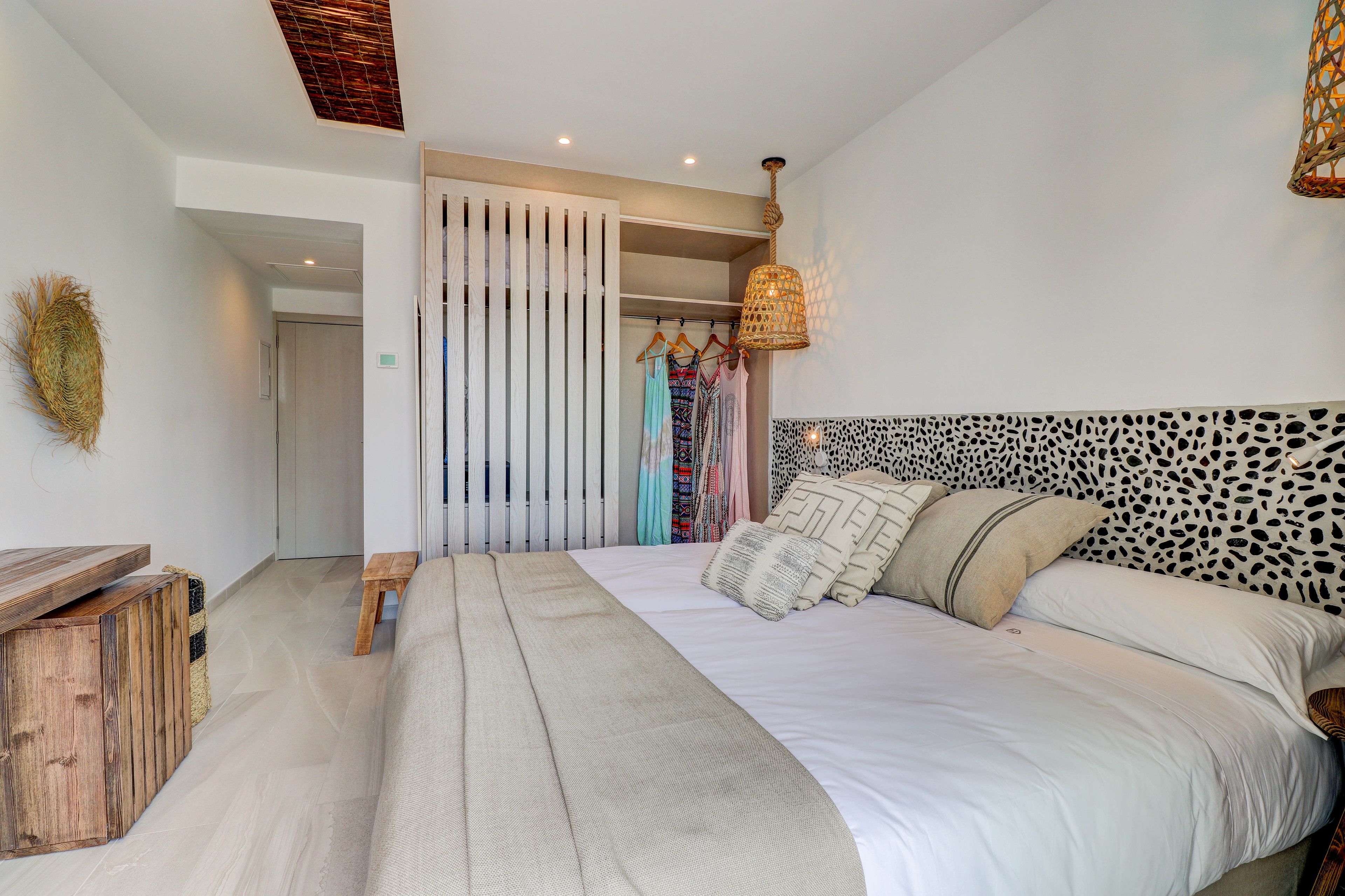 Ein Schlafzimmer mit einem großen Bett, einer hölzernen Bank, einer Pendelleuchte und einem Kleiderschrank mit Kleidung.