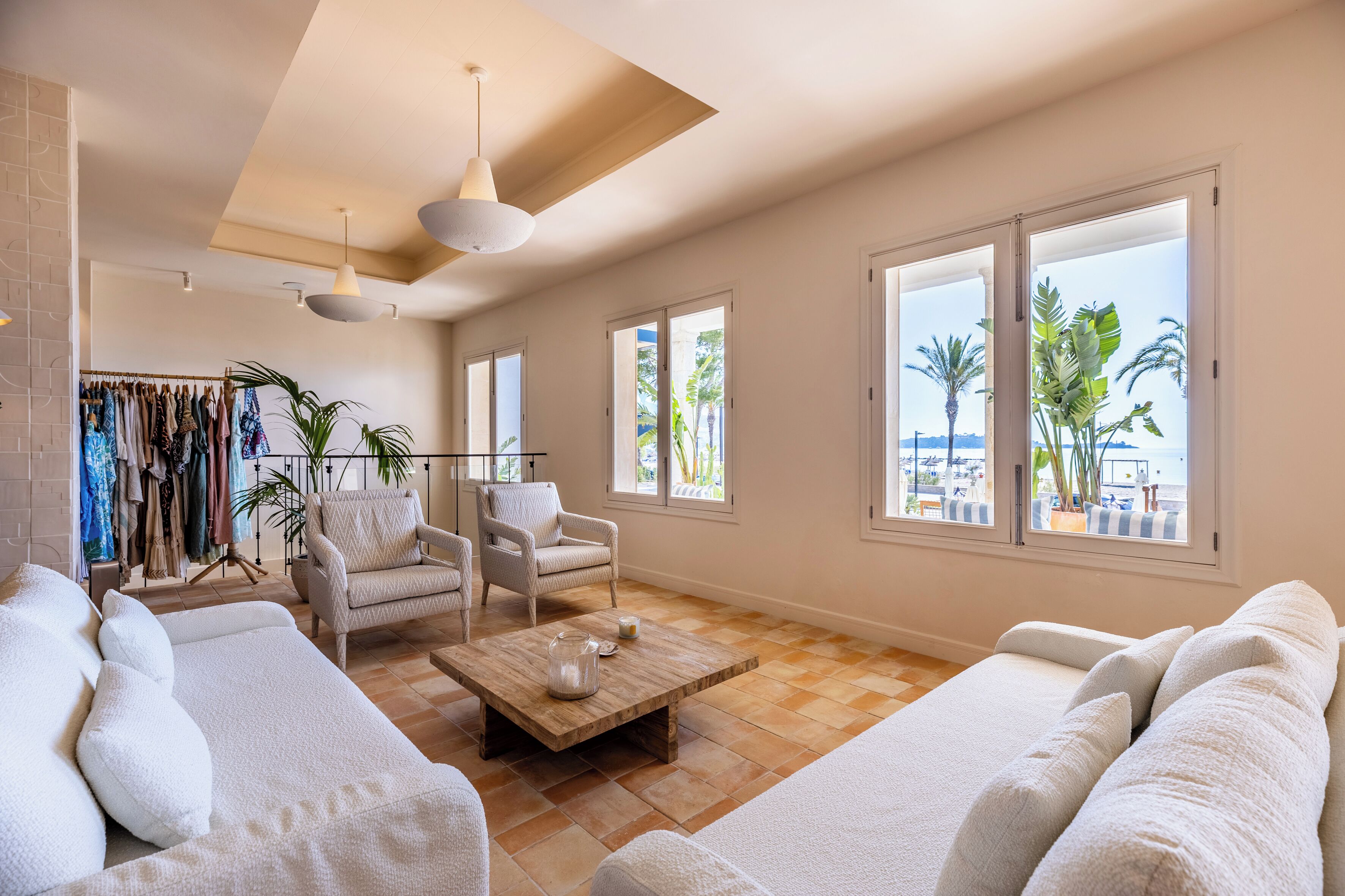 Un salon spacieux comprenant un ensemble de canapés, une table basse et un fauteuil. On y trouve également un portant à vêtements avec divers articles et une plante en pot. De grandes fenêtres offrent une vue sur des palmiers et une plage.
