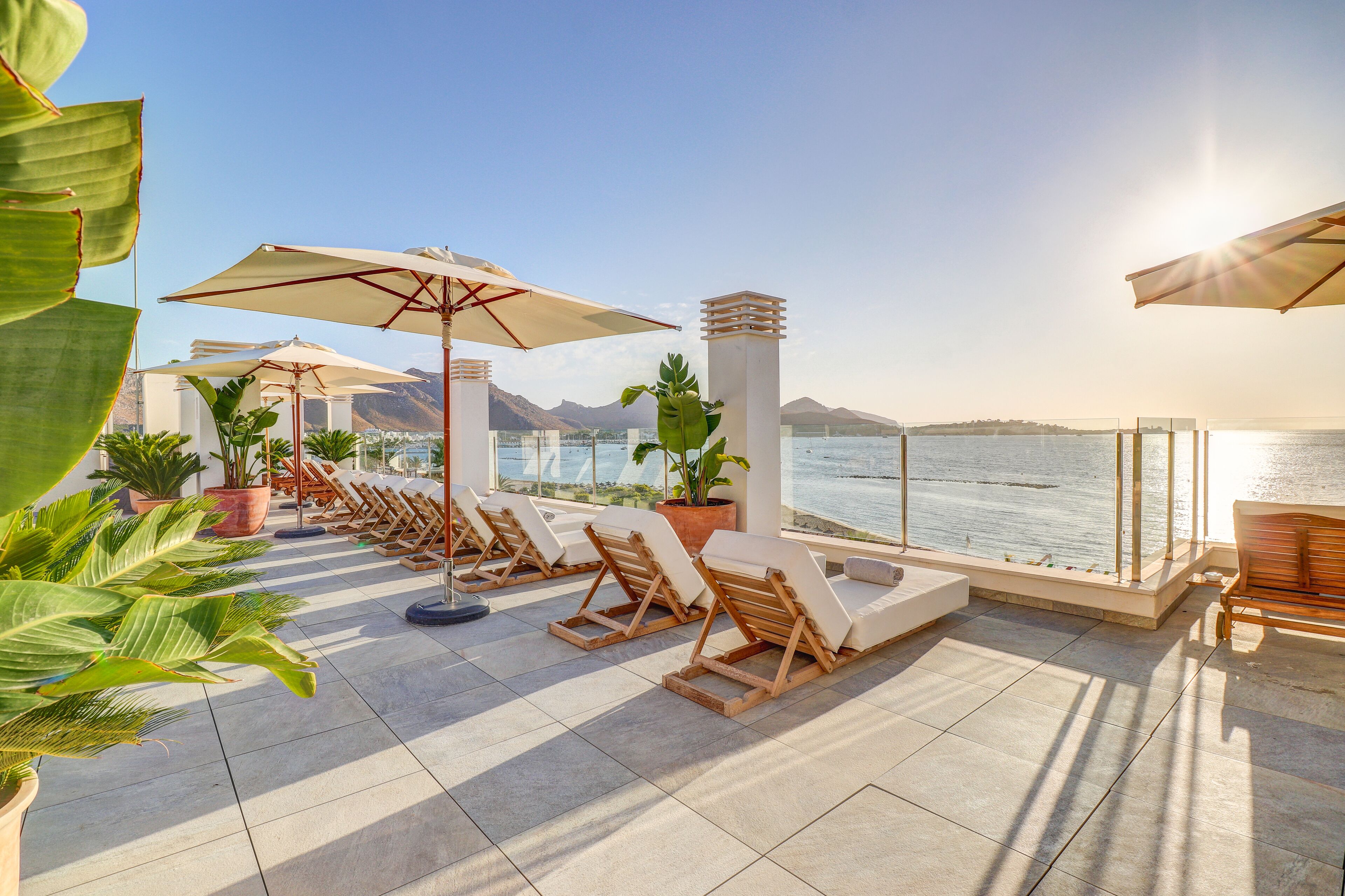 A sunlit terrace with lounge chairs and umbrellas overlooking a body of water and mountains.