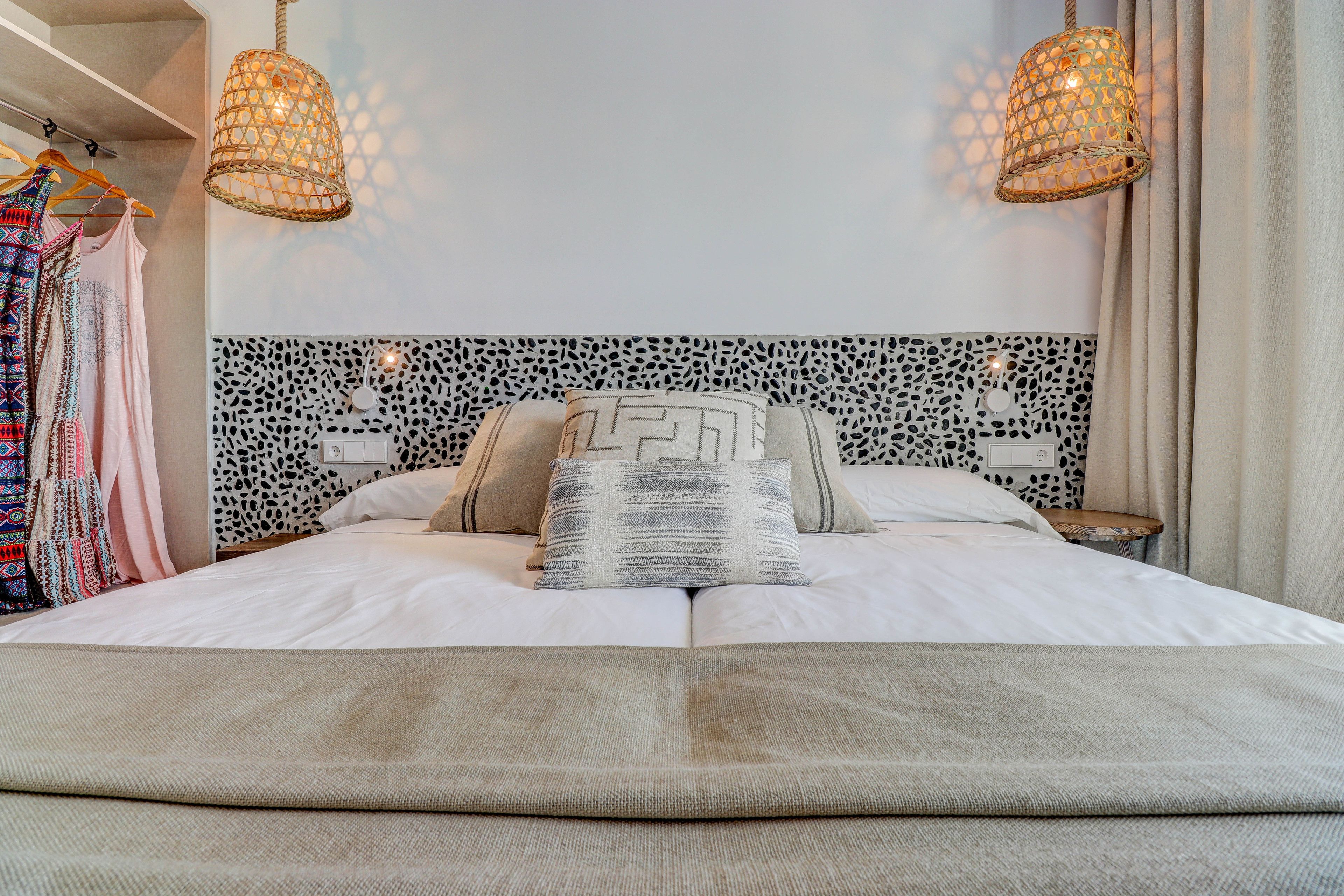 Una cama bien tendida con almohadones estampados, cabecera con diseño geométrico blanco y negro, y dos lámparas colgantes.