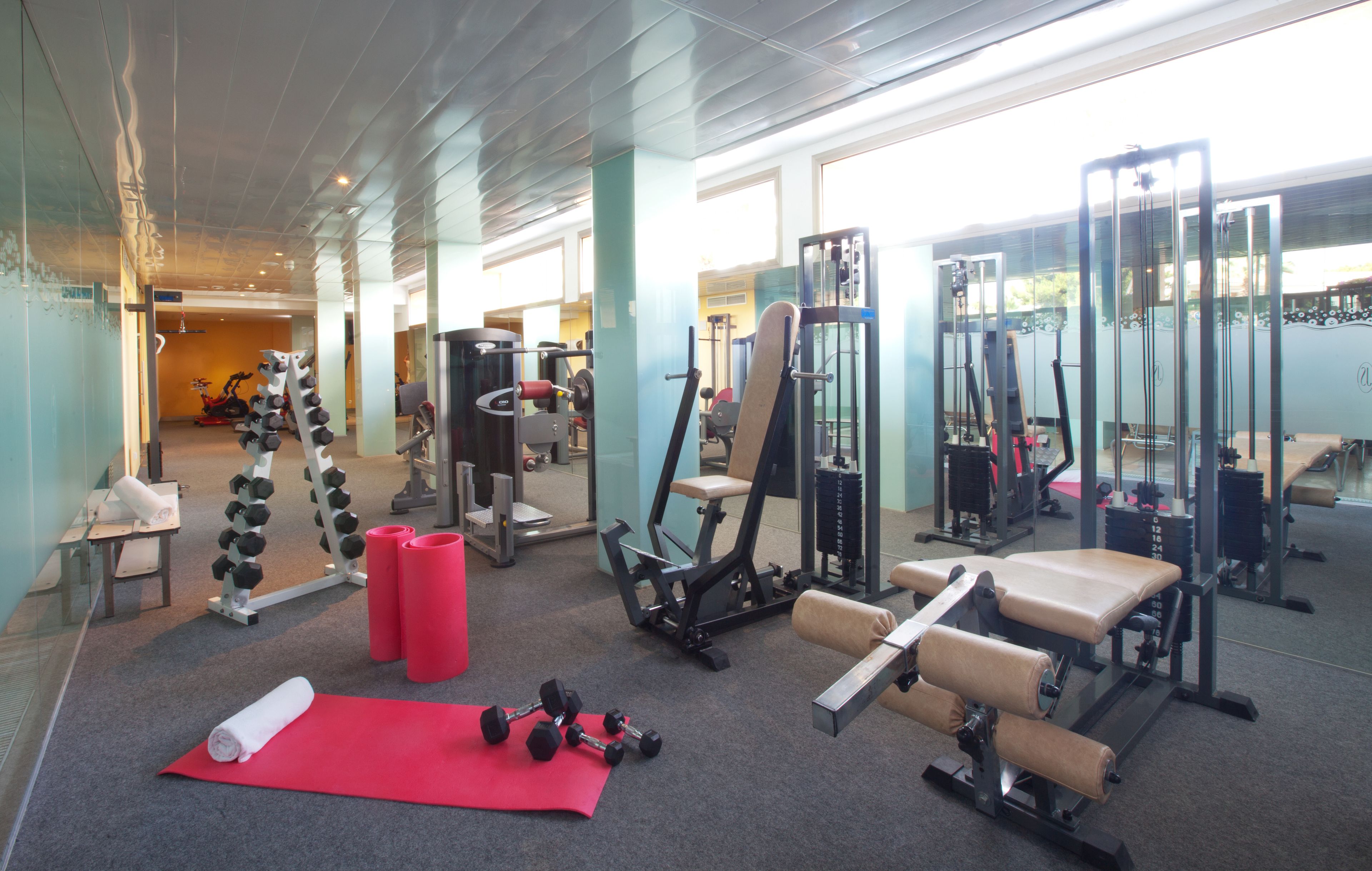 Un gimnasio con diversos equipos de ejercicio, una colchoneta roja y mancuernas.