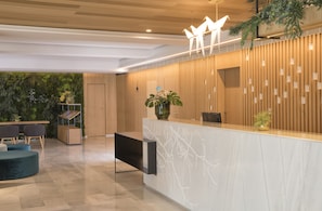A modern reception area with a wooden reception desk, a seating area with a round table and chairs, and a green wall with plants.