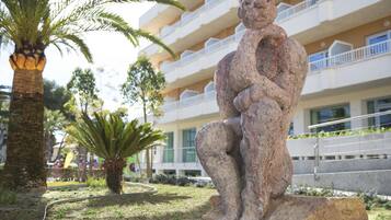 Una escultura de piedra de una figura sentada en un área ajardinada, con una palmera y un edificio al fondo.