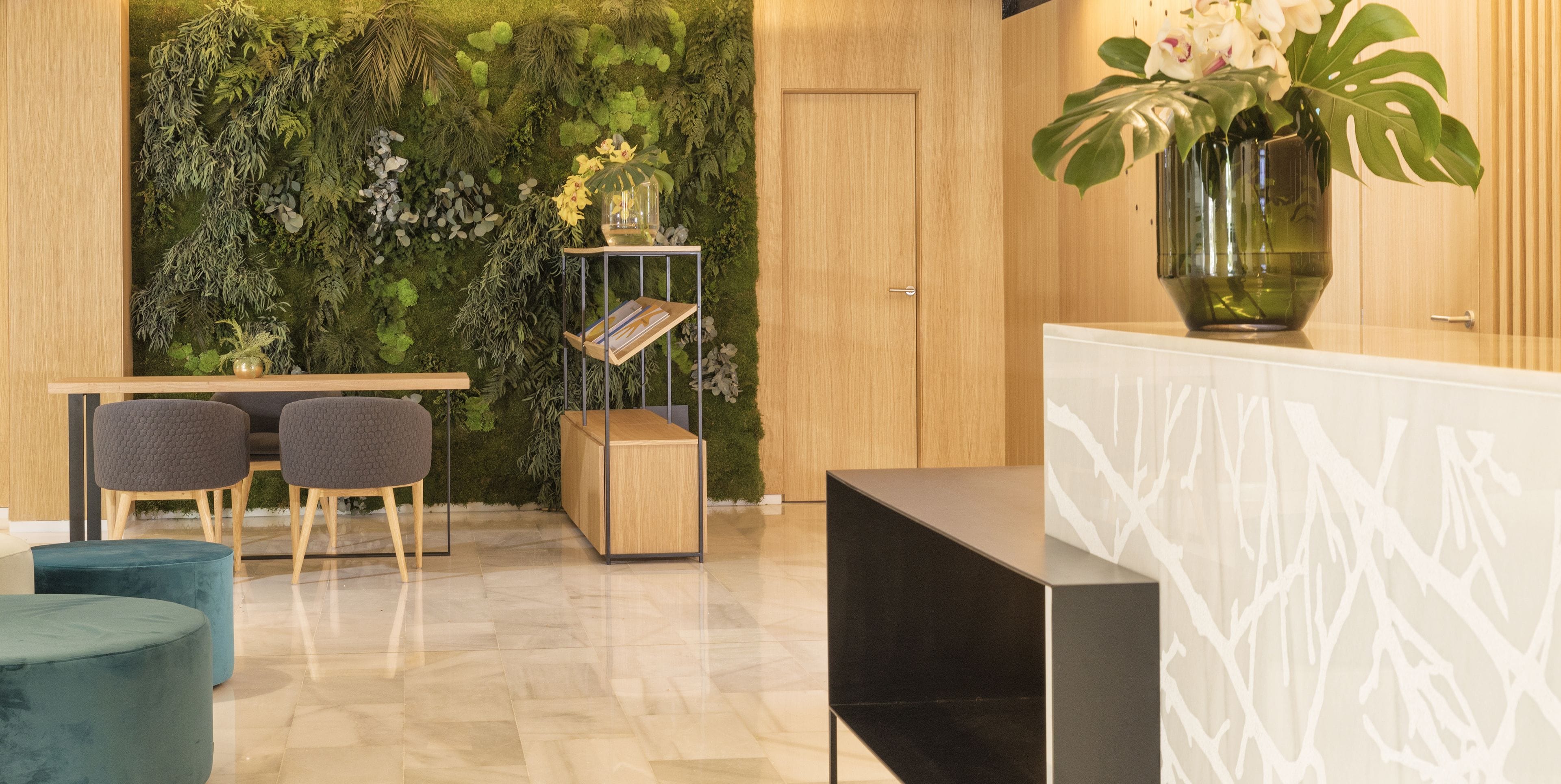 Een modern ingerichte receptie met een houten deur, een groene wand met planten, een receptiebalie, twee grijze stoelen en een blauwe poef.