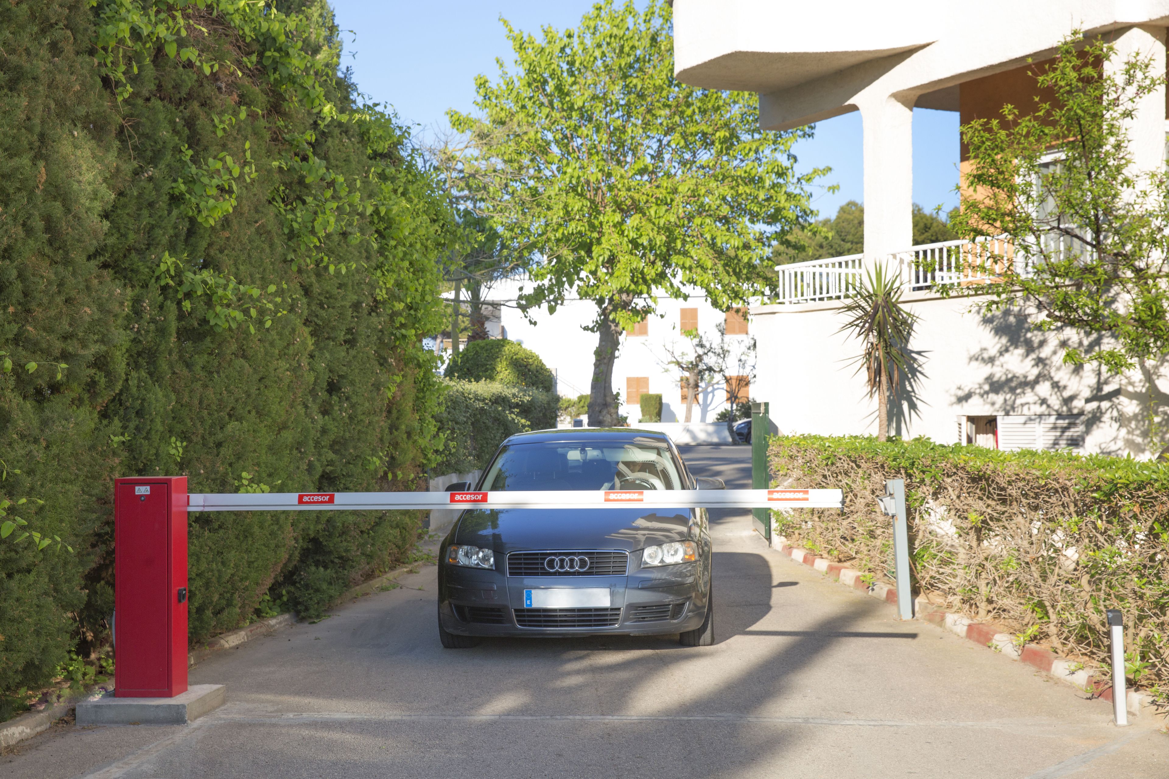 A gated entrance with a car parked inside, a red utility box, and a white building with balconies in the background.
