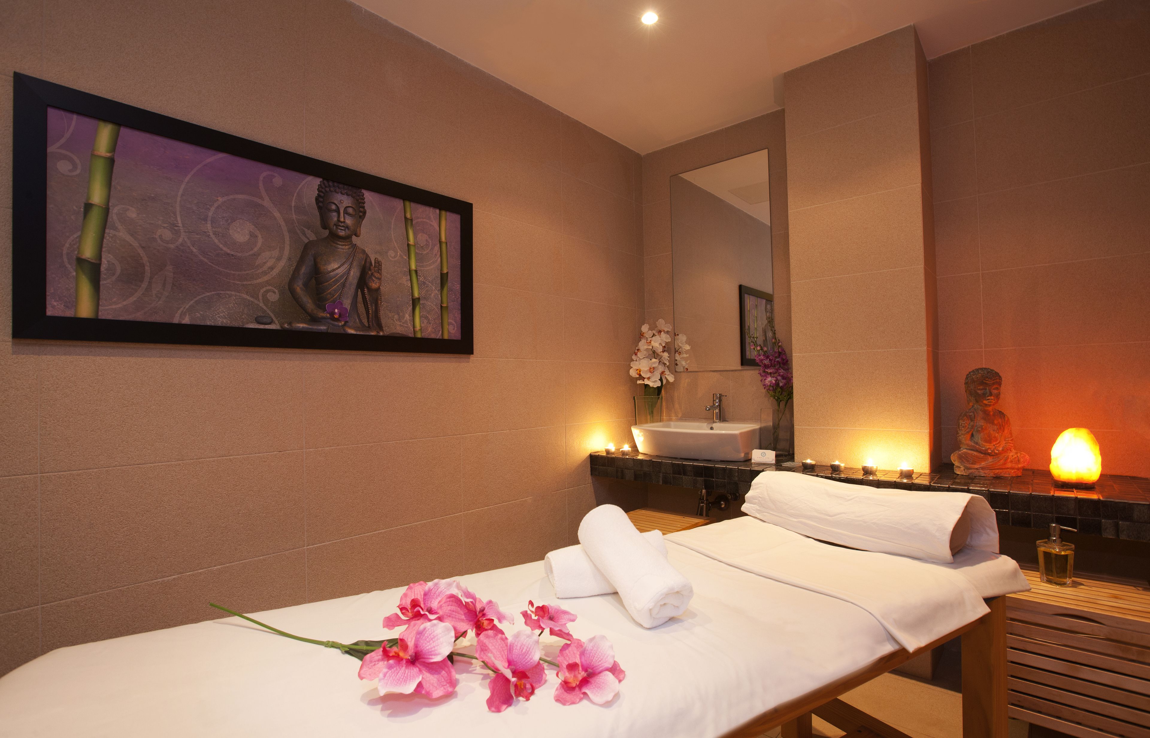 Ein Spa-Raum mit Massagetisch, Buddha-Statue, Spiegel, Waschbecken und Kerzen.