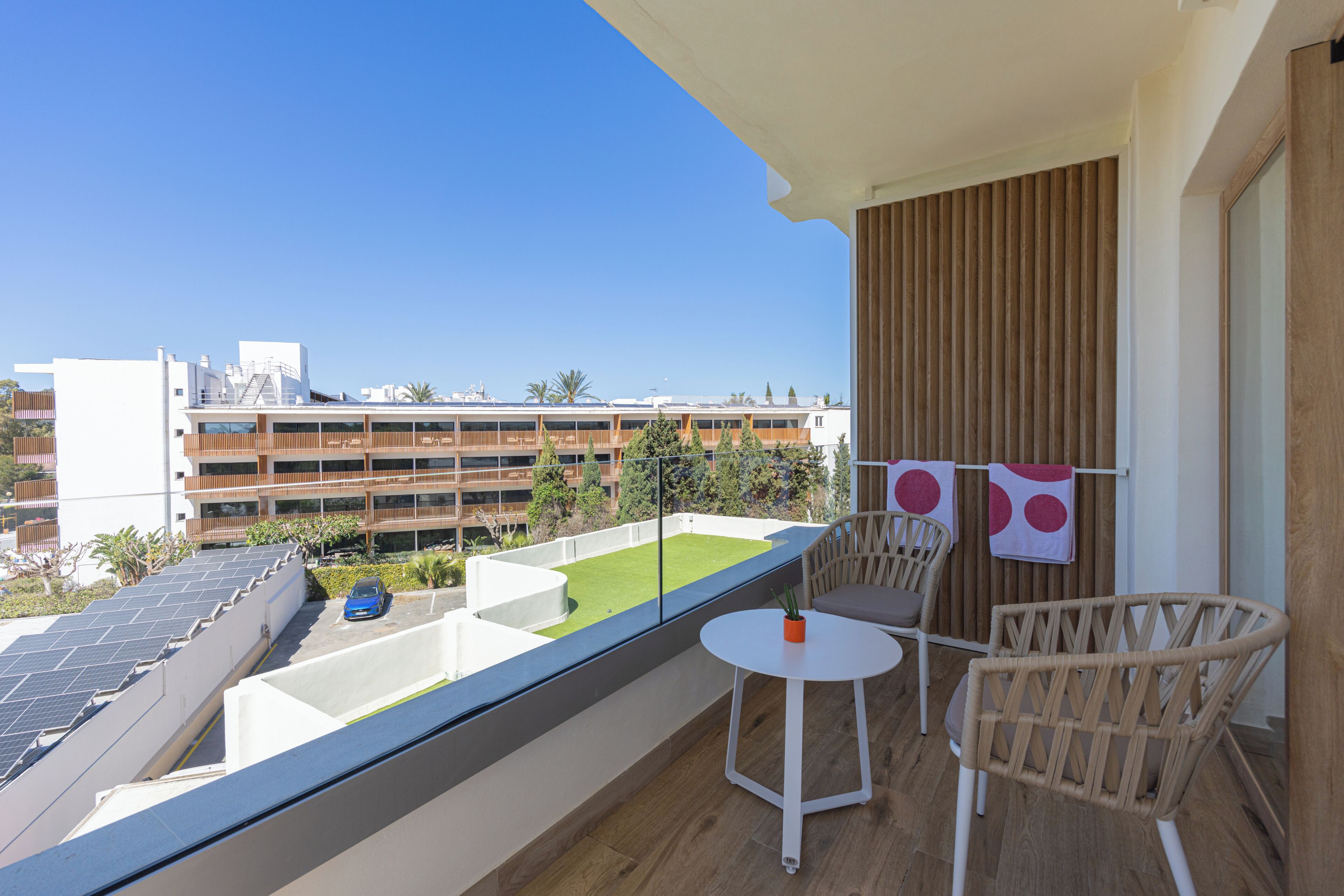A balcony with a view of a building complex, a small table, two chairs, and a blue car parked outside.