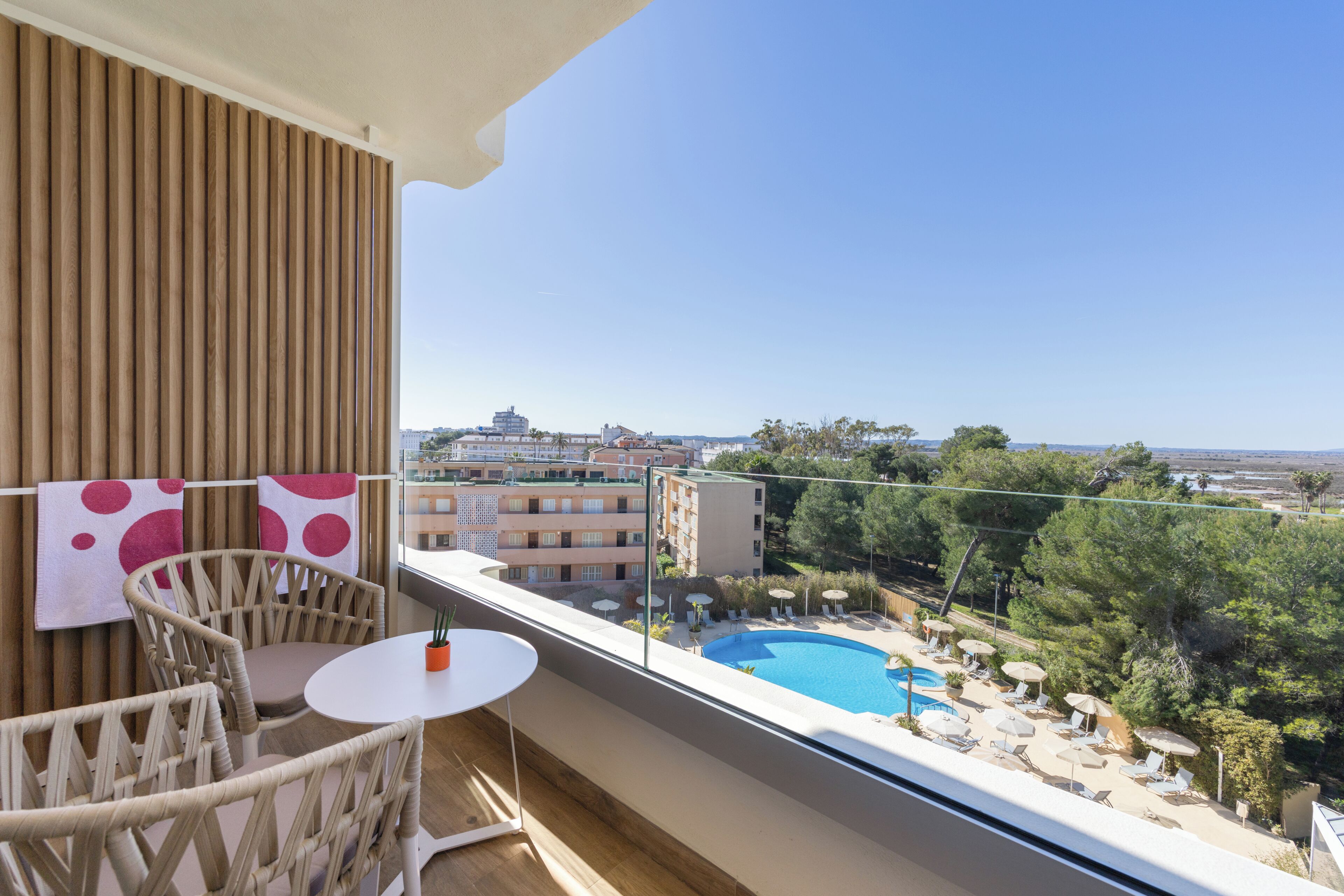 A balcony with wicker chairs, a small table, and a view of a swimming pool and surrounding buildings.