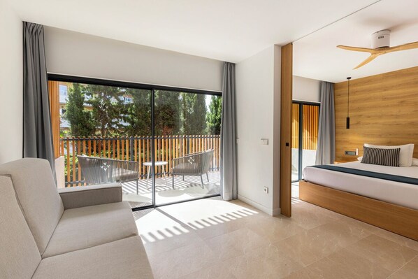 A modern hotel room with a large sliding glass door to a balcony, a sofa, a bed with a wooden headboard, and a ceiling fan. - Las Gaviotas Suites Hotel (Muro)